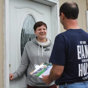 A woman stands smiling, holding the doorknob to her front door. A man faces her, back to the camera, holding a clipboard with literature on it.