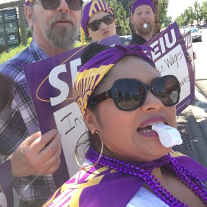 A group of SEIU members wear purple and wave signs on a picket line. A woman in the foreground prepares to blow on a plastic whistle.