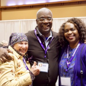 Three people standing together with arms around each others' shoulders, smiling, wearing purple SEIU lanyards, in a conference hall