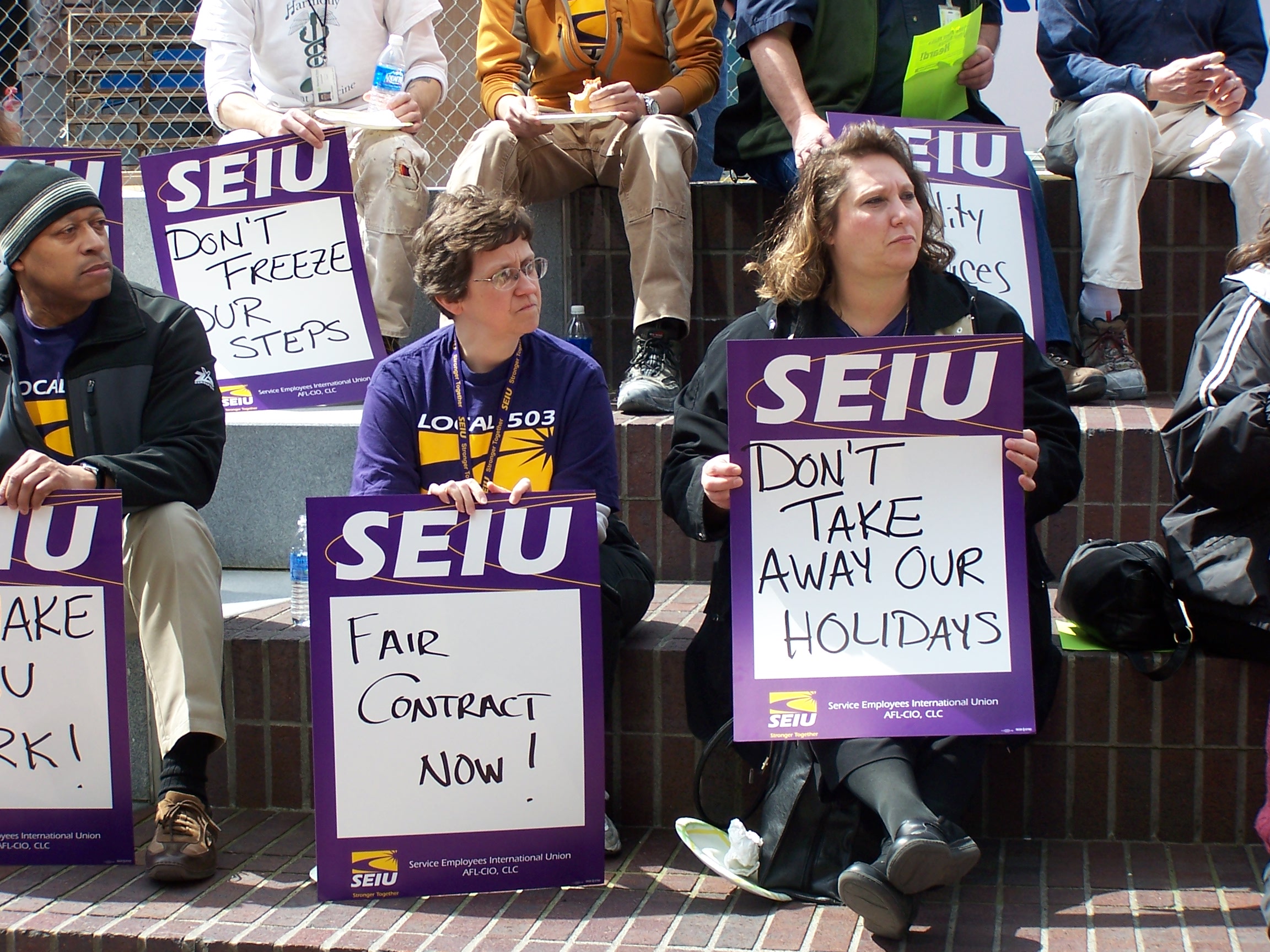 Một nhóm các thành viên SEIU và những người ủng hộ ngồi trên các bậc thang bằng gạch, lắng nghe ai đó nói. Họ cầm những tấm biển ghi dòng chữ "Hợp đồng công bằng ngay!" và "Đừng làm mất đi kỳ nghỉ của chúng tôi"