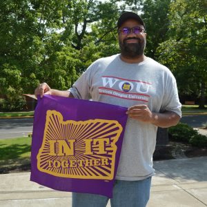 An SEIU member stands against a green field of trees, holding a purple and yellow bandana up that reads "IN IT TOGETHER"