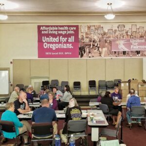 Группа членов профсоюза на собрании в большом конференц-зале. На стене позади них висит баннер с надписью «Едины для всех жителей штата Орегон» и изображением марширующих членов.