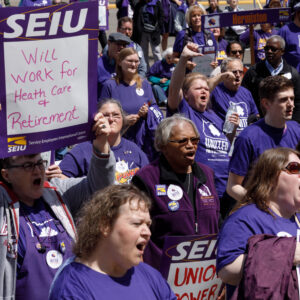 толпа людей в фиолетовых футболках SEIU, один из которых держит табличку с надписью «Будет работать в сфере здравоохранения и пенсионного обеспечения».