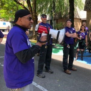 An SEIU member leader in a purple shirt speaking to coworkers on a megaphone. They are on a shady street and there are sunny trees and buildings behind them.