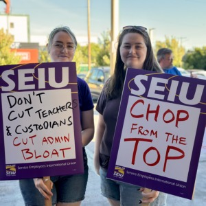 Two PPS custodians holding SEIU signs. One says "Don't cut teachers & custodians, cut admin bloat". The other says "Chop from the top"
