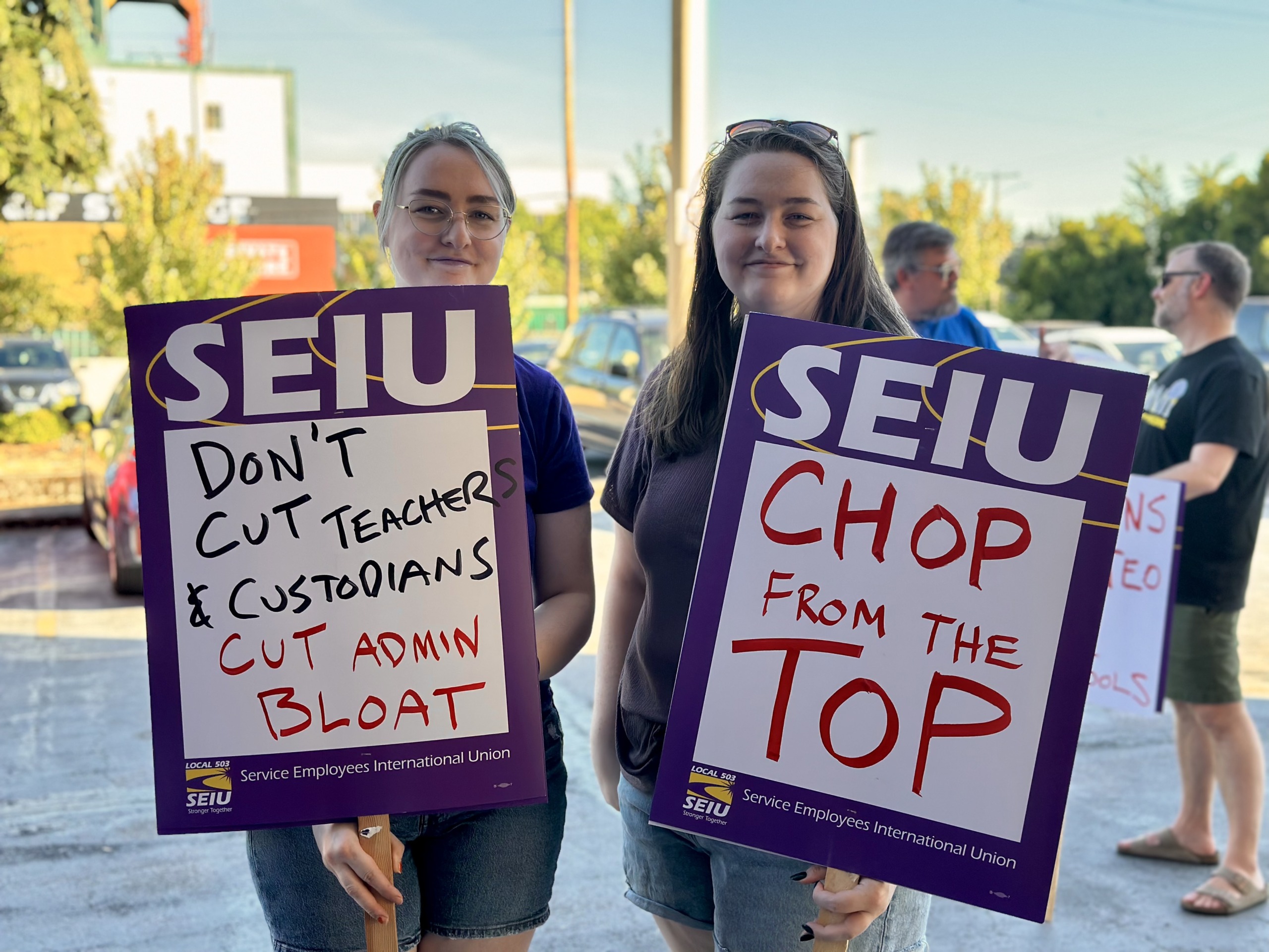 Two PPS custodians holding SEIU signs. One says "Don't cut teachers & custodians, cut admin bloat". The other says "Chop from the top"