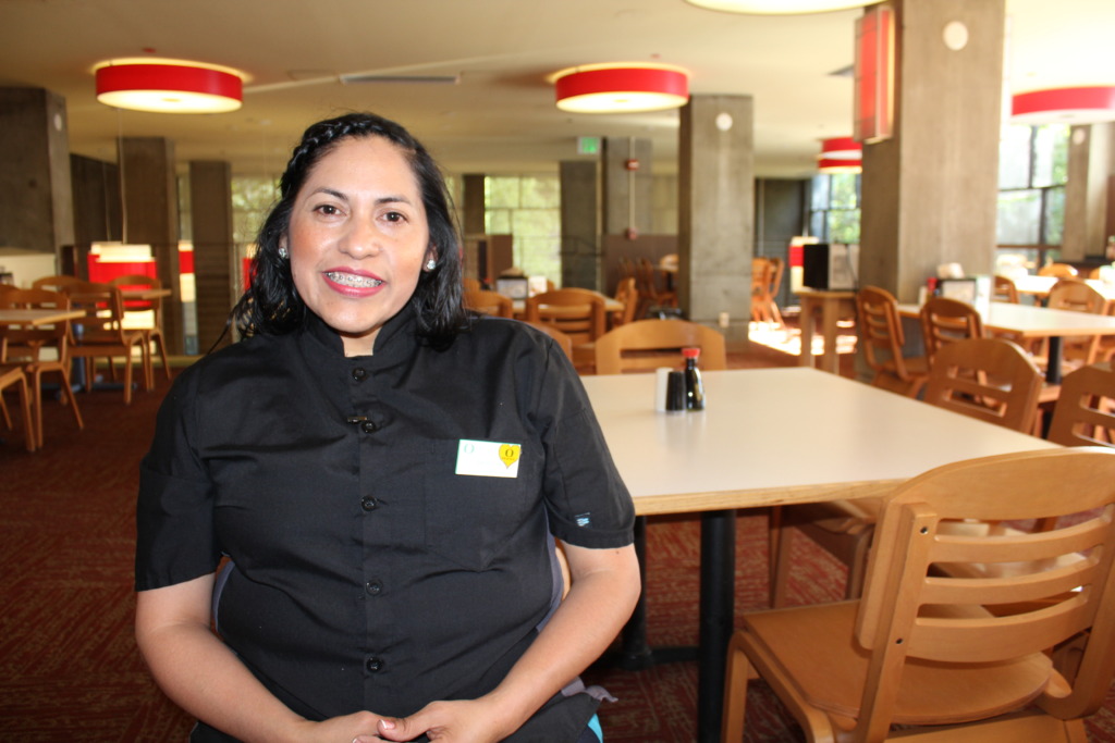 Una mujer sonriente vistiendo un uniforme de servicio de comidas universitario, en una cafetería universitaria vacía