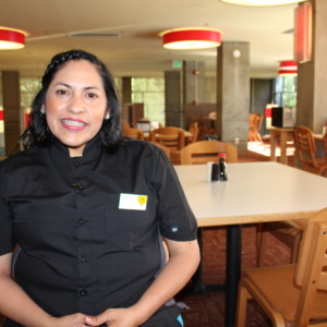 Một người phụ nữ mỉm cười mặc đồng phục dịch vụ ăn uống của trường đại học, trong căn tin trống của trường đại học