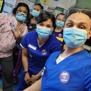 A group of nursing home workers wearing scrubs, surgical masks, and purple SEIU stickers, in a nursing home staff break room