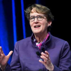 A woman with short hair and glasses wearing a purple jacket, speaking in front of a blue curtain