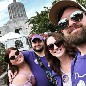 Un grupo de miembros del sindicato con gafas de sol, algunos con camisetas moradas de SEIU, parados al aire libre, abrazados y sonriendo. El edificio del Capitolio del Estado de Oregón está al fondo.