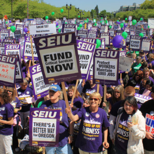 Большая толпа членов SEIU стоит на улице в солнечный день в фиолетовых футболках и с табличками SEIU. Некоторые говорят: «Финансируйте критически важные службы сейчас», «Лучший путь для Орегона» и «Мы едины».