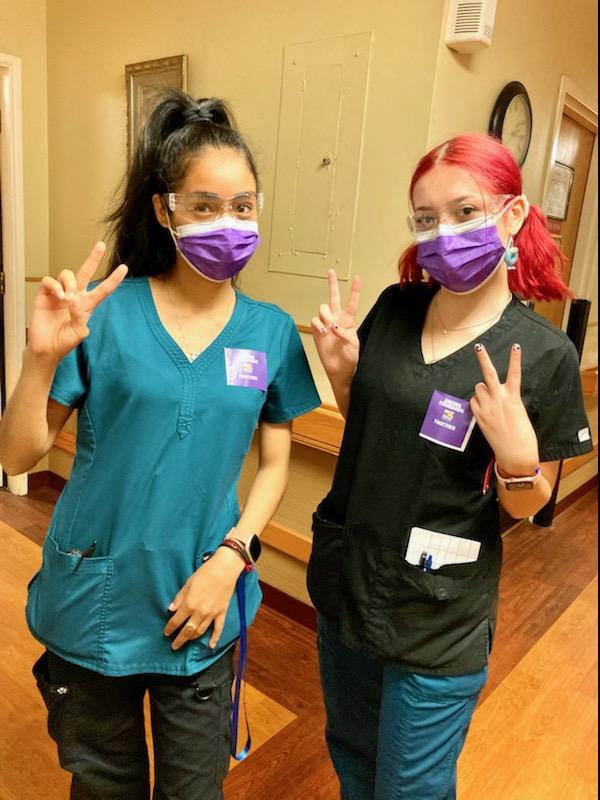 Two young women wearing purple surgical masks and scrubs, holding up two fingers in the 'victory' sign. One has dark hair and one has hair dyed pink. They are standing in a nursing home.