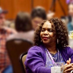 A woman wearing a purple SEIU sweatshirt sitting at a table at a conference, smiling and looking off to the left. There are blurred images of a crowd of people behind her