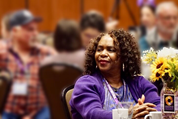 Una mujer con una sudadera morada de SEIU sentada en una mesa en una conferencia, sonriendo y mirando hacia la izquierda. Hay imágenes borrosas de una multitud de personas detrás de ella.