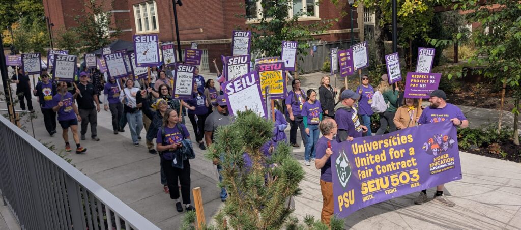 A group of workers marching in purple t shirts carrying banners and signs: Universities united for a fair contract. Background is a brick building on the PSU campus
